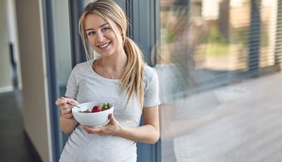 Junge schwangere Frau isst eine Schale mit Erdbeeren und schaut sehr freundlich.