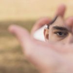 Eine Hand, die eine Spiegelscherbe hält, in der ein Auge zu sehen ist.