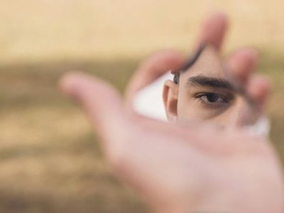 Eine Hand, die eine Spiegelscherbe hält, in der ein Auge zu sehen ist.