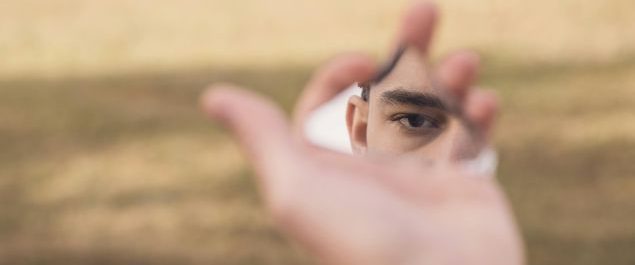 Eine Hand, die eine Spiegelscherbe hält, in der ein Auge zu sehen ist.